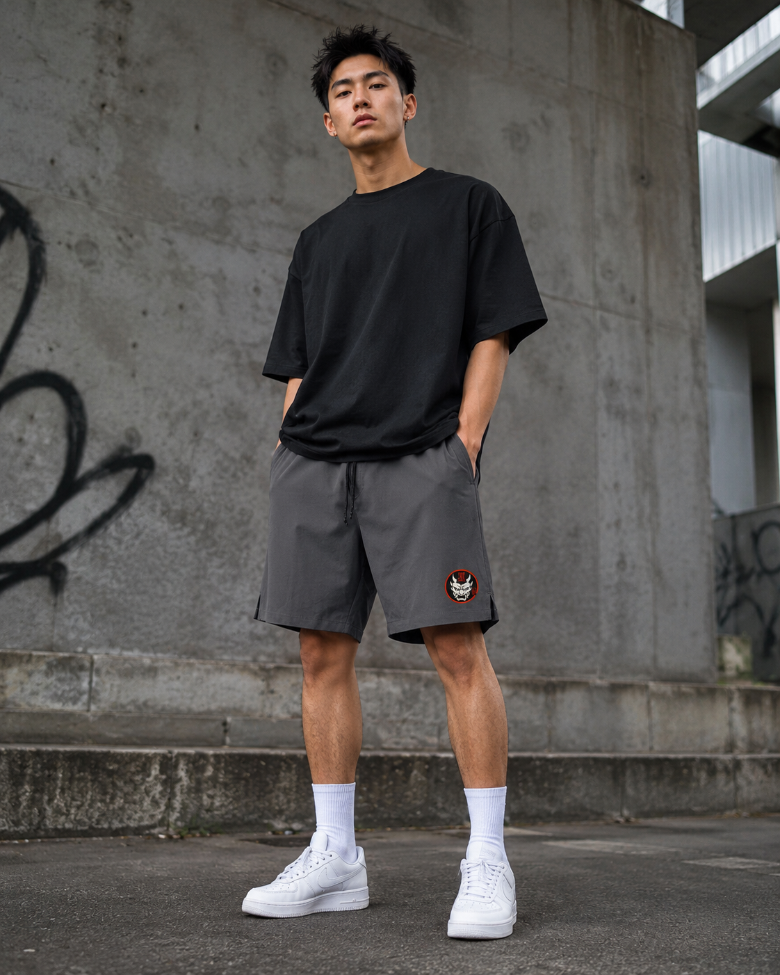 A young man stands outdoors by a graffiti wall, hands in his pockets, wearing loose black irezumi ink clothing—a T-shirt and Kuro Oni Streetwear Shorts with an embroidered patch—paired with white socks and trainers.